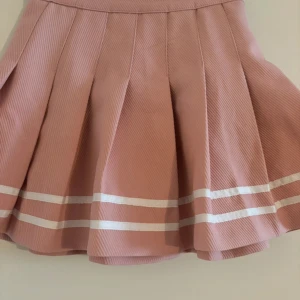 Rosa kjol från H&M Divided - Superfin rosa kjol från H&M Divided i storlek 34. Den är gjord av polyester och har en söt design med vita ränder längst ner. Perfekt för vår och sommar! Kjolen har en normal passform och är i mycket bra skick. Har ett litet veck (se sista bilden) men inget man tänker på eller som syns så mycket. Köparen betalar frakt. 