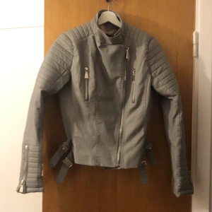 Grå bikerjacka från Voyelles - Säljer en snygg grå bikerjacka från Voyelles i storlek 42. Jackan har coola dragkedjor och bältesdetaljer, perfekt för höst och vår. Materialet är polyuretan med polyesterfoder, vilket ger en skön känsla. Passar perfekt för en edgy look!
