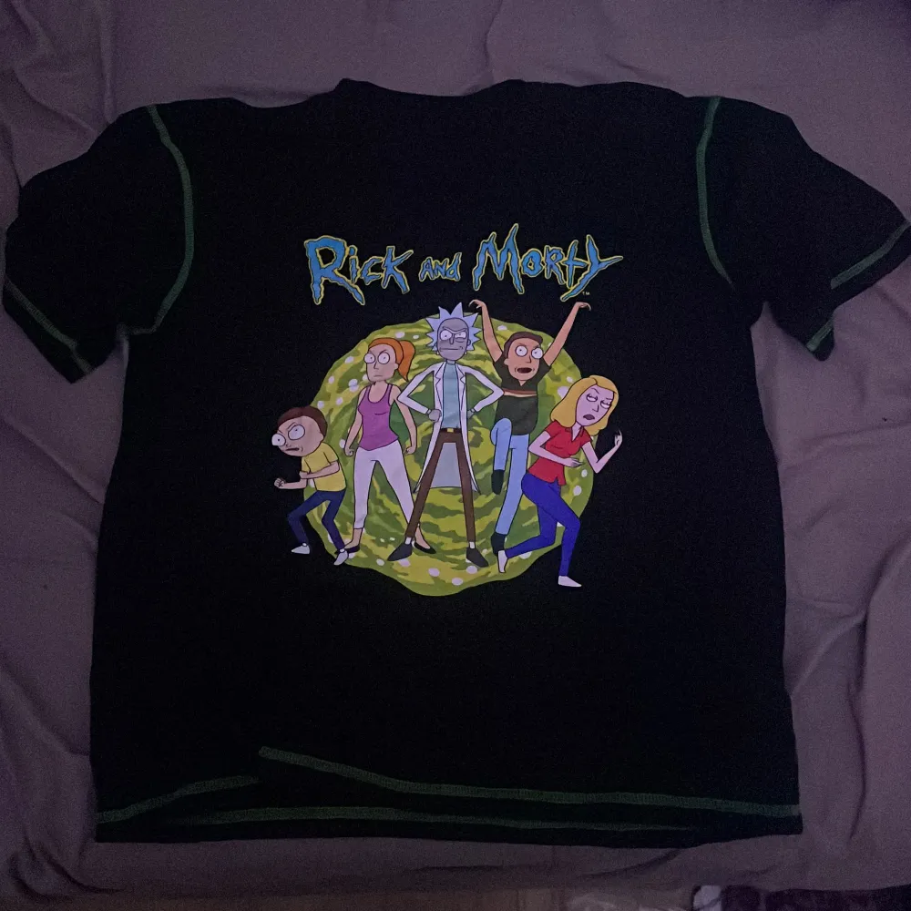 Cool svart t-shirt med Rick and Morty-tryck på framsidan. Perfekt för fans av serien! T-shirten har korta ärmar och är i bra skick. Den är gjord av bomull och har gröna detaljer vid ärmslut och hals. Perfekt för en avslappnad stil eller att bära på en fest med vänner.. T-shirts.