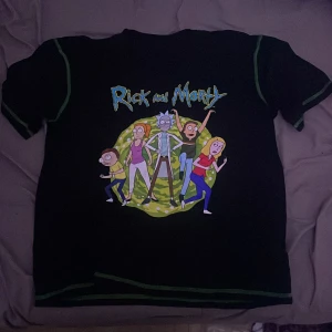 Svart Rick and Morty t-shirt - Cool svart t-shirt med Rick and Morty-tryck på framsidan. Perfekt för fans av serien! T-shirten har korta ärmar och är i bra skick. Den är gjord av bomull och har gröna detaljer vid ärmslut och hals. Perfekt för en avslappnad stil eller att bära på en fest med vänner.
