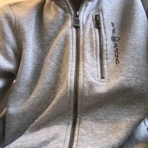 Grå hoodie från Sail Racing - Säljer en snygg grå hoodie från Sail Racing i mycket bra skick. Den har en dragkedja framtill och en praktisk bröstficka med dragkedja. Perfekt för både vardag och träning. Passar bra till höst och vår!