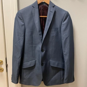 Blå kavaj från Valentino - Säljer en elegant blå kavaj från Valentino i Super 150 ull. Kavajen har en klassisk design med två knappar och fickor fram. Insidan är fodrad med ett lyxigt mönster i rött. Perfekt för både formella och semi-formella tillfällen.