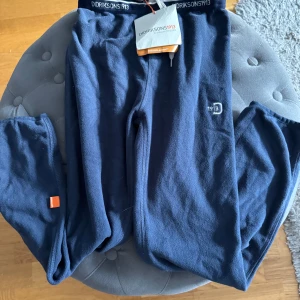 Nya: Mörkblå fleecebyxor från Didriksons stl 130 - Säljer ett par mörkblå fleecebyxor från Didriksons i nytt skick, lappar på. . Byxorna har en elastisk midja med märkets logga och en liten orange detalj vid benet. Superbekväma och stilrena för både inomhus och utomhus.