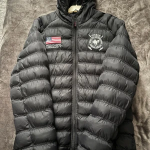 Svart pufferjacka från Vinson Polo Club - Säljer en svart pufferjacka från Vinson Polo Club i mycket bra skick. Jackan har en broderad logga och dragkedja framtill. Perfekt för kyliga dagar med sin quiltade design och huva. Passar både till vardags och lite finare tillfällen.