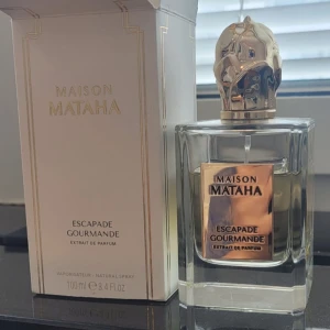 Maison Mataha Escapade Gourmande - Säljer en lyxig parfym från Maison Mataha, Escapade Gourmande. Flaskan är elegant med en fyrkantig form och en unik guldfärgad kork som ser ut som en skulptur. Parfymen är 100 ml och kommer i en stilren vit förpackning. Perfekt för den som älskar exklusiva dofter!