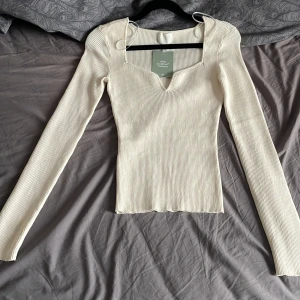 Vit ribbad topp från H&M - Säljer en superfin vit ribbad topp från H&M. Den har en unik v-ringad design och långa ärmar, perfekt för både vardag och fest. Materialet är mjukt och stretchigt, vilket gör den bekväm att bära hela dagen. Passar perfekt till jeans eller kjol! Androg använd och är i väldigt fina skick.