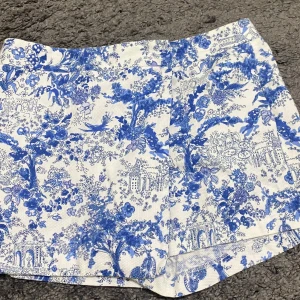 Blåvita blommiga shorts från ASOS💙🤍 - Säljer ett par superfina blåvita shorts från ASOS med ett detaljerat blommönster. Perfekta för sommardagar eller en avslappnad vårlook. De är i mycket bra skick och har en normal passform. Materialet känns lätt och bekvämt, perfekt för varma dagar! Liknar kjol men är shorts 💙🤍