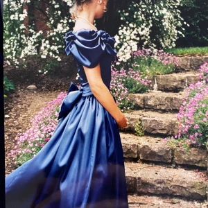 Balklänning gråblå - Säljer min mammas balklänning från 95 i perfekt skick, endast använd två ggr.Elegant mörkblå långklänning i siden med puffiga kortärmar, och en stor rosett därbak (avtagbar). Klänningen har en klassisk och tidlös design som ger en kunglig känsla. Klänningen är specialsydd för tjej som är 1.69-1.70 cm lång (storlek uppskattas att va 34). Finns en innerkjol (se separat bild) som är avtagbar. Ett riktigt kap då den är både vacker som den är, men även lätt att göra om. Rekommenderas ej för stor byst