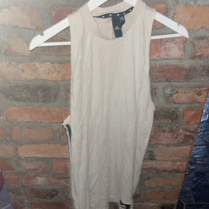 Beige ärmlös klänning från Adidas - Säljer en snygg beige ärmlös klänning från Adidas i storlek S. Perfekt för träning eller en avslappnad dag. Den har de klassiska Adidas-ränderna på sidan och är gjord i ett mjukt och bekvämt material. Den är något genomskinlig så tips att ha något under typ cyckel byxor 