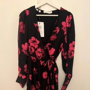 Svart blommig omlottklänning från Selected Femme - Snygg svart omlottklänning med rosa blommor från Selected Femme. Klänningen har långa ärmar och ett knytband i midjan för en smickrande passform. Perfekt för både fest och vardag! Nypris 1000kr aldrig använd!