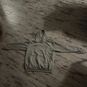 Grå hoodie från Gina Tricot - Säljer en snygg grå hoodie från Gina Tricot med texten 'MA CHÉRIE' i glittriga detaljer på framsidan. Perfekt för en casual look eller mysiga dagar hemma. Den har en klassisk passform med långa ärmar och en bekväm huva. Passar perfekt till jeans eller leggings!