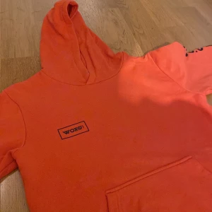 Orange hoodie med svart text vid armarna och vid bröstet - Säljer en cool orange hoodie med tryck på bröstet och ärmen. Den har en stor ficka fram och en mysig huva. Perfekt för höst och vår när du vill vara både bekväm och stilren. Passar bra till jeans eller joggers!