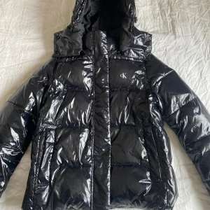 Svart pufferjacka från Calvin Klein - Säljer en snygg svart pufferjacka från Calvin Klein i mycket bra skick. Jackan har en glansig yta och är perfekt för kalla dagar. Den har en dragkedja framtill och en praktisk huva. Perfekt för vintern och hösten!