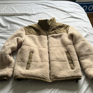 The North Face fleecejacka - Säljer en supermysig beige teddyjacka från The North Face. Den har en cool kontrast med olivgröna detaljer på axlarna och dragkedjorna. Perfekt för kyliga dagar med sin fluffiga och varma design. Perfekt för vintern. Vid snabb affär 950kr
