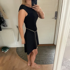 Svart klänning från Dancing Days - Säljer en elegant svart klänning från Dancing Days. Den har en snygg kedjedetalj i midjan och är ärmlös med en stilren design. Perfekt för festliga tillfällen! Klänningen har en dragkedja på sidan för enkel påklädning.