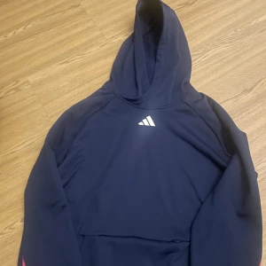 Adidas Tröja  - En Grymt mörkblå huvtröja med gula och rosa ränder på sidan av armarna. Tröjan har en ficka på magen, en huva & en logotyp på bröstet. Skick 10/10 då den inte använts mer än 3 gånger