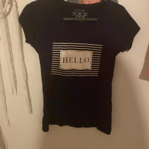 Svart t-shirt med 'HELLO'-tryck - Säljer en svart t-shirt från Divided med ett coolt 'HELLO'-tryck i vitt och ränder runtom. Den är kortärmad och perfekt för en avslappnad stil. Passar bra till jeans eller shorts för en casual look.