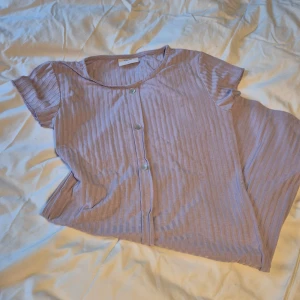 Ljuslila ribbad topp från Grunt - Säljer en superfin ljuslila ribbad T-shirt klänning från Grunt i storlek L. Den har korta ärmar och knappar framtill, vilket ger en söt och avslappnad look. Perfekt för vår och sommar!
