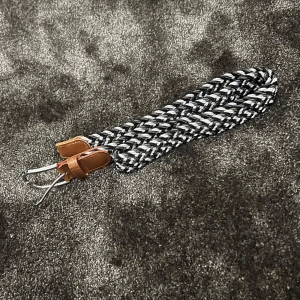 Flätat bälte med läderdetaljer - Säljer ett snyggt flätat bälte i svart och vitt med bruna läderdetaljer. Bältet har en klassisk design med ett silverfärgat spänne. Perfekt för att ge en stilren touch till vilken outfit som helst!