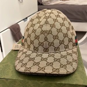Beige och brun keps från Gucci - Säljer en snygg keps från Gucci med deras ikoniska GG-mönster i beige och brunt. Kepsen har en justerbar rem baktill i brunt skinn och klassiska gröna och röda detaljer på sidorna. Perfekt för att ge din outfit en lyxig touch!
