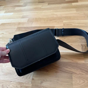 Svart axelväska med dragkedja - Säljer en stilren svart axelväska i skinn. Väskan har en praktisk dragkedja och flera fack för enkel organisering. Perfekt för både vardag och fest!