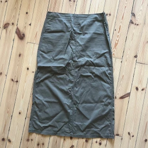 Olivgrön långkjol - Säljer en stilren olivgrön långkjol i ett mjukt och bekvämt material. Kjolen har en klassisk design med två fickor bak och en diskret slits för extra rörelsefrihet. Perfekt för både vardag och festliga tillfällen!