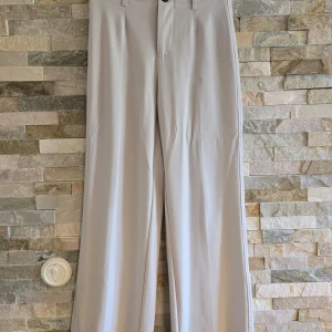 Beige byxor från Bershka - Säljer ett par snygga beige byxor från Bershka. De är högmidjade och har en stilren design med vida ben. Perfekta för både vardag och fest! Byxorna har en knapp och dragkedja framtill för enkel på- och avtagning.