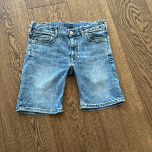 Jeansshorts i blå denim - Säljer ett par snygga jeansshorts i blå denim. De har en klassisk femficksdesign med en liten logga på den lilla fickan fram. Shortsen har en knappgylf och är i en bekväm längd som slutar strax ovanför knäna. Perfekta för sommardagar!