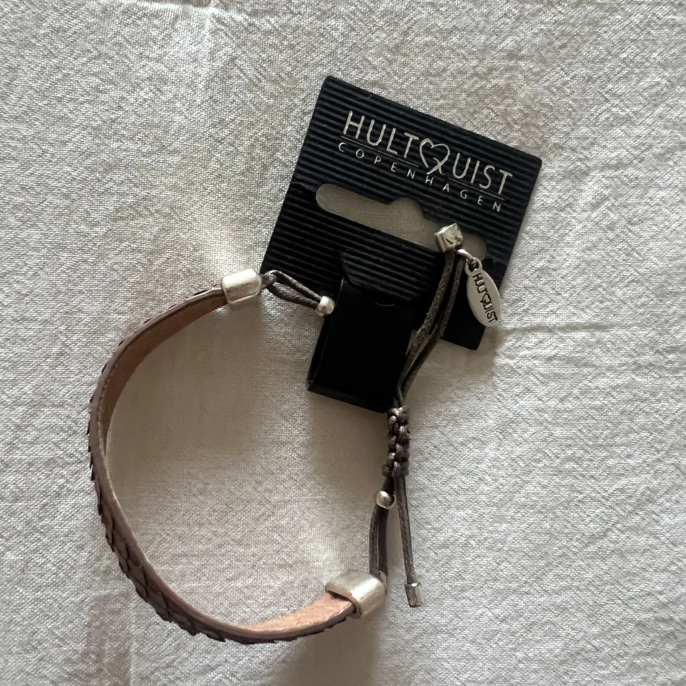 Snyggt brunt armband från Hultquist Copenhagen med en flätad design. Armbandet har justerbara snören och silverfärgade detaljer. Perfekt accessoar för både vardag och fest. Nytt och oanvänt med etikett kvar. Helt nytt!. Accessoarer.