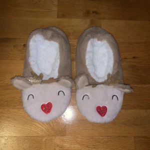  rentofflor  - Supermysiga tofflor i beige med gulliga renansikten och röda hjärtan på nosen. De har vita fluffiga insidor och glittriga horn. Perfekta för att hålla fötterna varma under vintern och julen. Sulan är prickig för extra grepp. Bara testade en gång så som nya!
