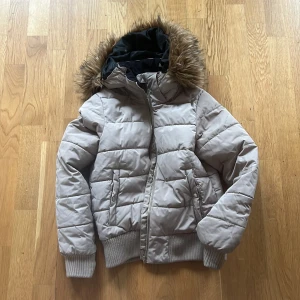 Beige pufferjacka med fuskpäls - Säljer en snygg y2k beige pufferjacka med fejkpäls på huvan. Jackan har dragkedja och ribbade muddar vid ärmslut och nederkant. 