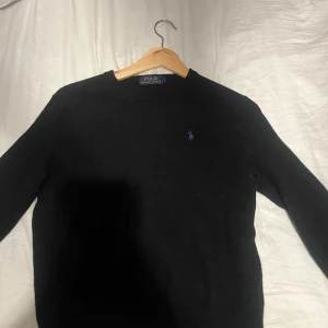 Svart tröja från Polo Ralph Lauren - Säljer en klassisk svart tröja från Polo Ralph Lauren med deras ikoniska  lila logga på bröstet. Tröjan är i bra skick och perfekt för både vardag och lite finare tillfällen. Den har en enkel och stilren design med rund hals och långa ärmar.