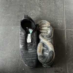 Svarta Nike Vapormax - Säljer ett par svarta Nike Vapormax i bra skick. De har en cool design med en stickad ovandel och en transparent sula. Skorna har en unik snörningslösning med en låsmekanism. Perfekta för både träning och vardagsbruk!