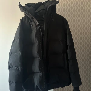 Svart pufferjacka från Canada Goose macmillan - Säljer en svart pufferjacka från Canada Goose i mycket bra skick. Jackan har en varm och vadderad design med långärmade ärmar och en praktisk dragkedja framtill. Perfekt för kalla vinterdagar. Den har en snygg logga på ärmen och en justerbar huva för extra skydd mot väder och vind.