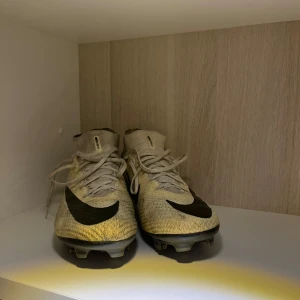Nike Mercurial superfly 9 elite fotbollsskor - Säljer ett par Nike Mercurial superfly 9 fotbollsskor i beige och svart. Skorna har en tight passform med snörning och är perfekta för fotboll. De är i bra skick och har en modern design med 'AIR' och 'ZOOM' detaljer på sidan. 