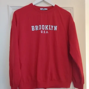 Röd sweatshirt med Brooklyn-tryck - Säljer en cool röd sweatshirt från Carla Mara med texten 'Brooklyn U.S.A.' i vitt på bröstet. Perfekt för en avslappnad stil och passar bra till jeans eller joggers. Tröjan har långa ärmar och en bekväm passform. Perfekt för höst och vår!