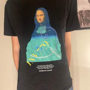 Cool svart t-shirt med ett färgglatt Mona Lisa-tryck från Musée du Louvre. Perfekt för konstälskare som vill ha en unik stil. T-shirten är kortärmad och har en normal passform. Passar bra till både vardag och fest!