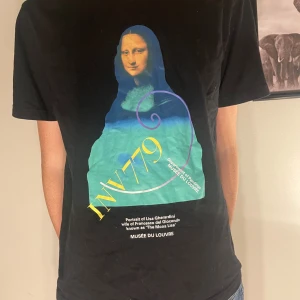 Svart t-shirt med Mona Lisa-tryck - Cool svart t-shirt med ett färgglatt Mona Lisa-tryck från Musée du Louvre. Perfekt för konstälskare som vill ha en unik stil. T-shirten är kortärmad och har en normal passform. Passar bra till både vardag och fest!