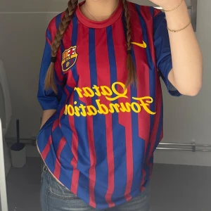 Blå och röd randig fcb fotbollströja - Säljer en klassisk fotbollströja från Nike i blått och rött med gula detaljer. Tröjan har korta ärmar och ett randigt mönster. Perfekt för fotbollsfans som vill visa sitt stöd. Skön och lätt att bära, passar både till match och vardag.