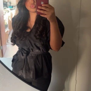 Svart blus med volangdetaljer - Säljer en elegant svart blus med volangdetaljer och knytband i midjan. Perfekt för både vardag och festliga tillfällen. Blusen har korta ärmar och en lös passform som ger en bekväm känsla. Passar bra till både jeans och kjol.