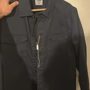 Mörkblå skjorta från SAMS - Säljer en stilren mörkblå skjorta från SAMS i storlek M. Skjortan har långa ärmar och två bröstfickor med lock. Perfekt för både vardag och fest! 🕺 Helt Ny!