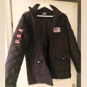 Svart quiltad jacka från Pogue - Säljer en svart quiltad jacka från Pogue i storlek M. Jackan har en dragkedja framtill och två fickor med knappar. Den har coola USA-detaljer på ärmen och bröstet. Perfekt för höst och vår!