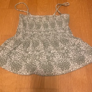 Blommig grön linne - Söt grönt linne med vita blommor och smala axelband. Perfekt för sommardagar! Den har en smockad överdel som ger en bekväm och stretchig passform. Passar bra till både jeans och kjol för en avslappnad look.