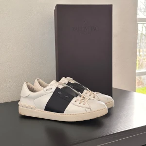 Valentino Garavani sneakers i vitt och navy - Säljer ett par snygga Valentino Garavani sneakers i vitt skinn med en bred navy rem över. Skorna har coola nitar på hälen och klassisk snörning. Perfekta för både vardag och fest! De är i bra skick med lite slitage på sulan. Kommer med originalförpackning.