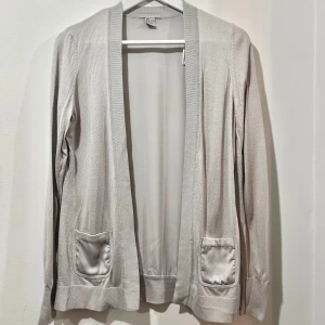 Grå kofta med fickor och silverderaljer - Säljer en stilren beige kofta i mycket bra skick. Den har långa ärmar och två praktiska fickor framtill. Perfekt för både vardag och lite finare tillfällen. Materialet känns mjukt och bekvämt mot huden. Passar perfekt till höst och vår!