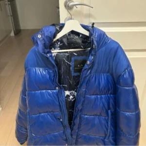 Blå pufferjacka från Armani Exchange - Säljer en snygg blå pufferjacka från Armani Exchange. Jackan har en glansig finish och är perfekt för kalla dagar. Den har en huva och stängs med dragkedja. Perfekt för både höst och vinter! Storlek L, fint skick på den. Passar både män och kvinnor/unisex. Finns i Örebro. Kan fraktas, köparen står för fraktkostnad.  Köpt på Boozt. 