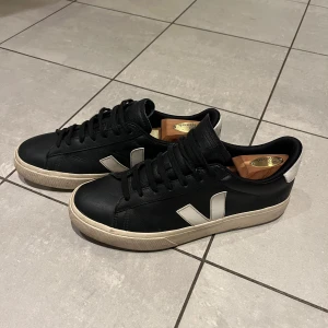 Svarta sneakers från Veja - Säljer ett par snygga svarta Veja sneakers i storlek 42. De har en vit sula och V-logga på sidan. Skorna är i bra skick med lite slitage på sulan. Perfekta för både vardag och fest!