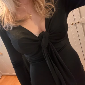 Svart klänning med knytning - Säljer en elegant svart klänning med v-ringning och knytning framtill. Den är långärmad och har en tight passform som framhäver figuren. Perfekt för festliga tillfällen eller en utekväll. Klänningen är i mycket bra skick och passar bra till både höst och vinter.