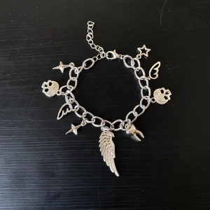 Handgjort Silverfärgat armband med berlocker - Coolt silverfärgat armband med olika berlocker som en vinge, stjärna, tand och dödskalle. ca 16 cm och kan justeras upp till 20 cm! Perfekt för att ge din outfit en edgy touch. Armbandet har en justerbar kedja för att passa olika storlekar. Passar både till vardags och festliga tillfällen!
