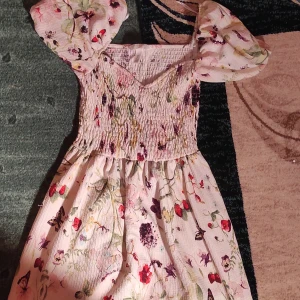 Blommig klänning med puffärmar - Superfin blommig klänning med puffiga kortärmar och smockdetaljer. Perfekt för sommarens fester eller en mysig picknick. Den har en söt v-ringning och är gjord i ett lätt och luftigt material. Färgerna är ljusa och somriga med inslag av rött, grönt och lila.